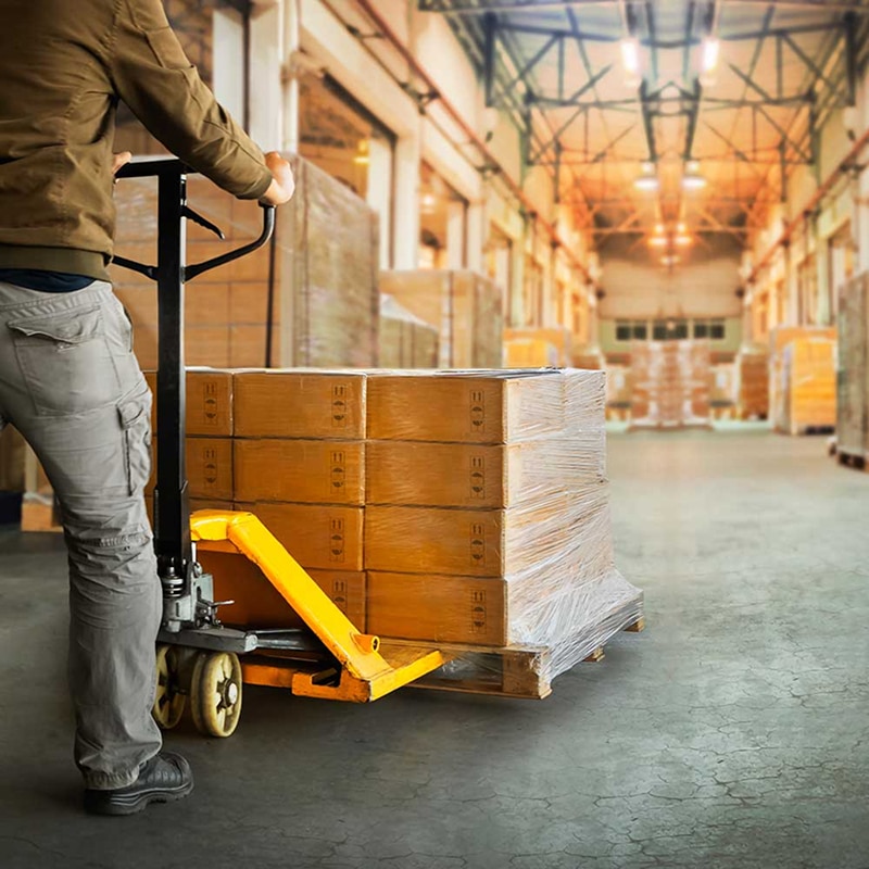 Manutentionnaire déplaçant des palettes de cartons emballés avec un transpalette manuel jaune dans un vaste entrepôt de logistique industriel.